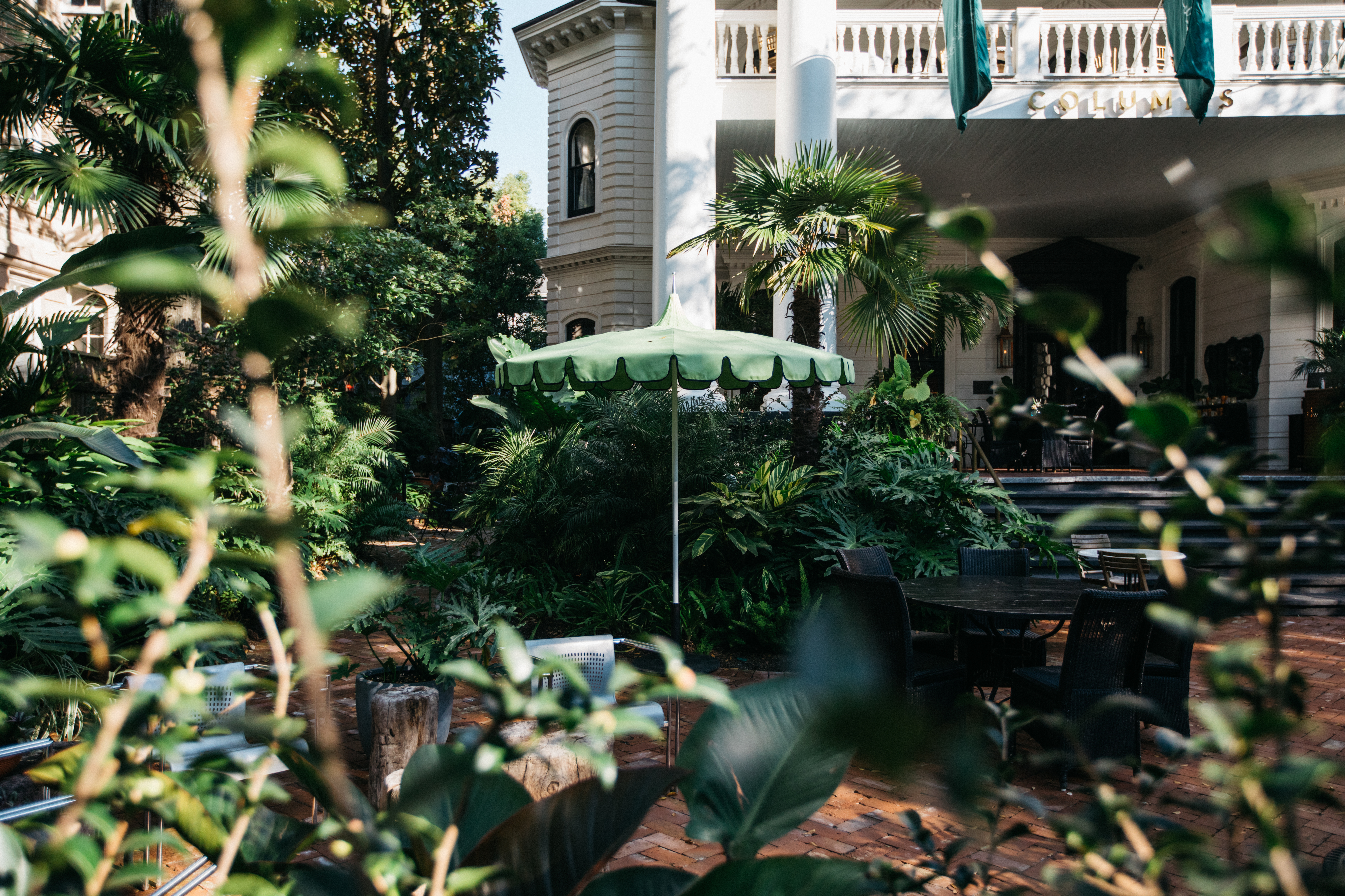 A courtyard with tables, chairs, and an umbrella surrounded by lush greenery and a balcony above.