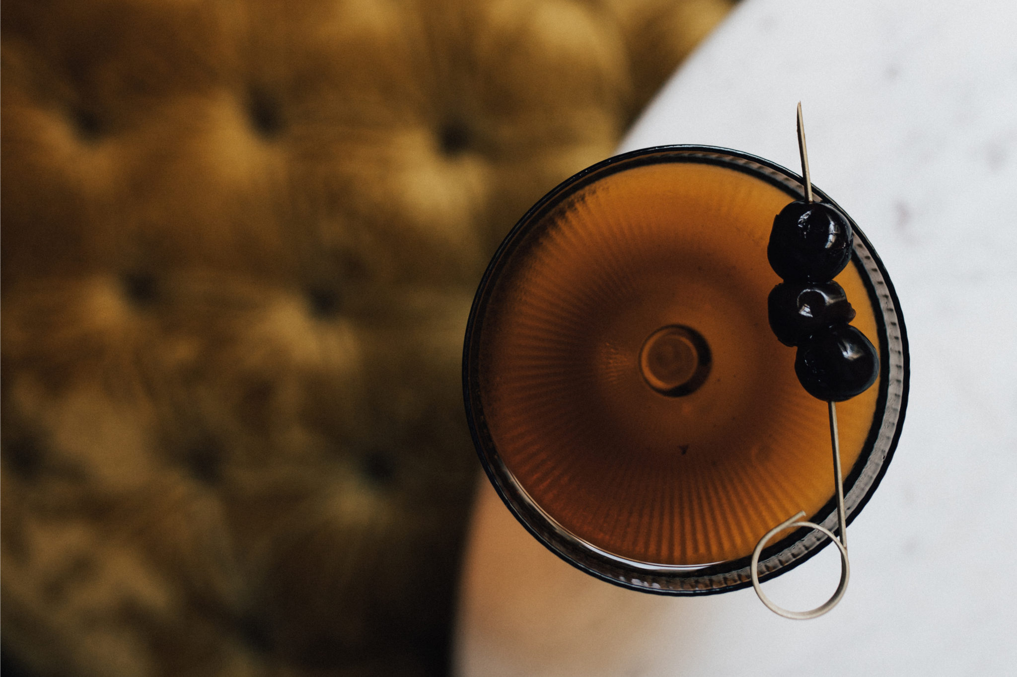 A cocktail glass with a brown drink and cherries on a table next to a tufted upholstered chair.