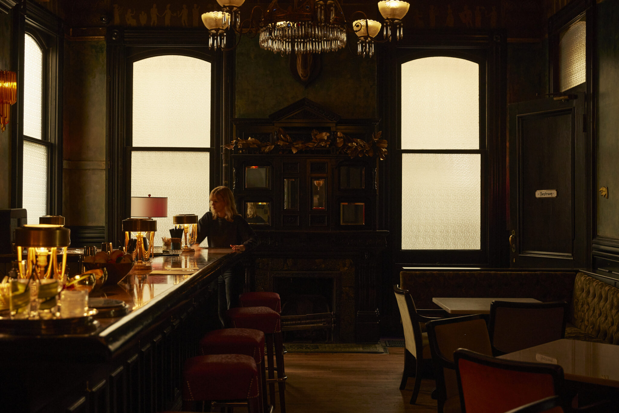 A dimly lit bar with a long counter, red stools, and a chandelier hanging from the ceiling.